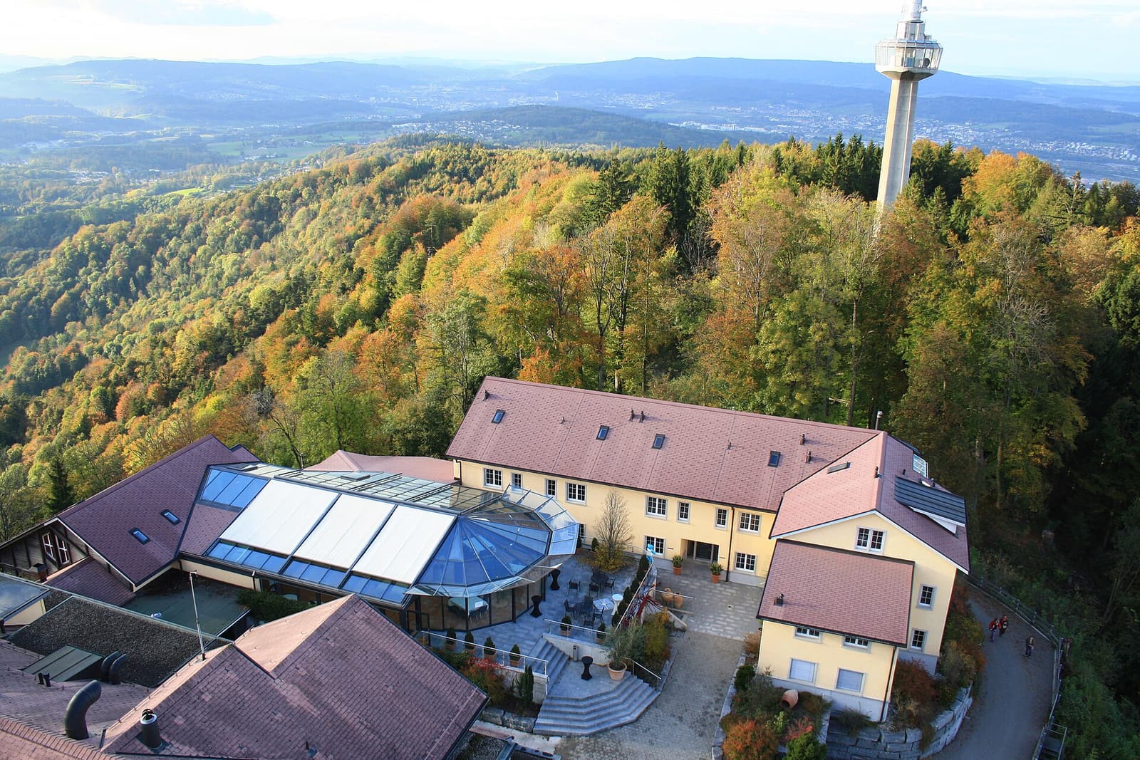 Les bâtiments de l’hôtel UTO KULM sur le sommet boisé vus d’en haut, avec la tour de télévision à l’arrière-plan